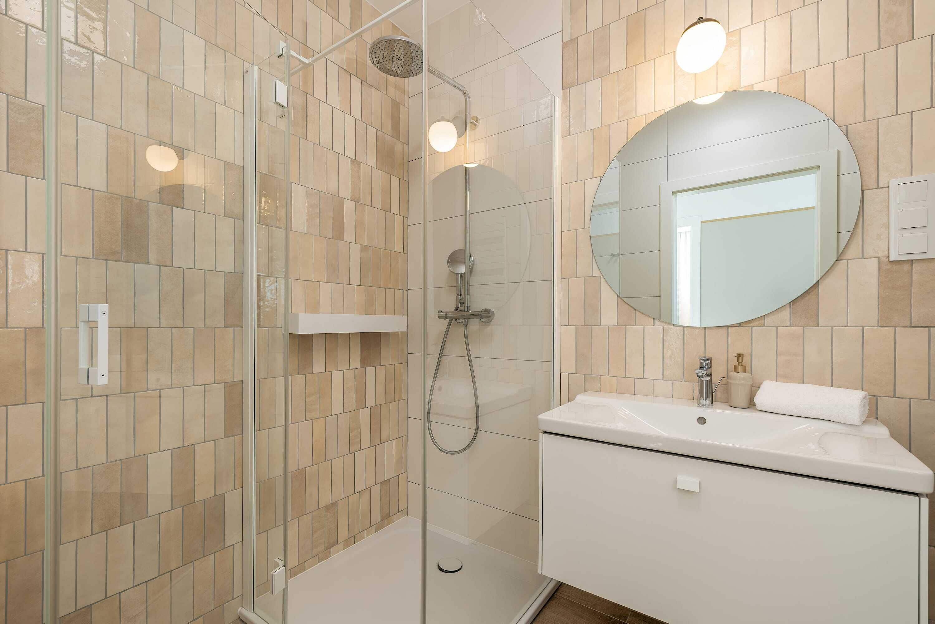 Un bany modern amb plat de dutxa de vidre, un mirall rodó i un lavabo blanc.