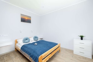 Une chambre avec un lit en bois, une couverture bleue, deux oreillers, une table de chevet, une commode blanche, une plante en pot et un tableau représentant un coucher de soleil accroché au mur.