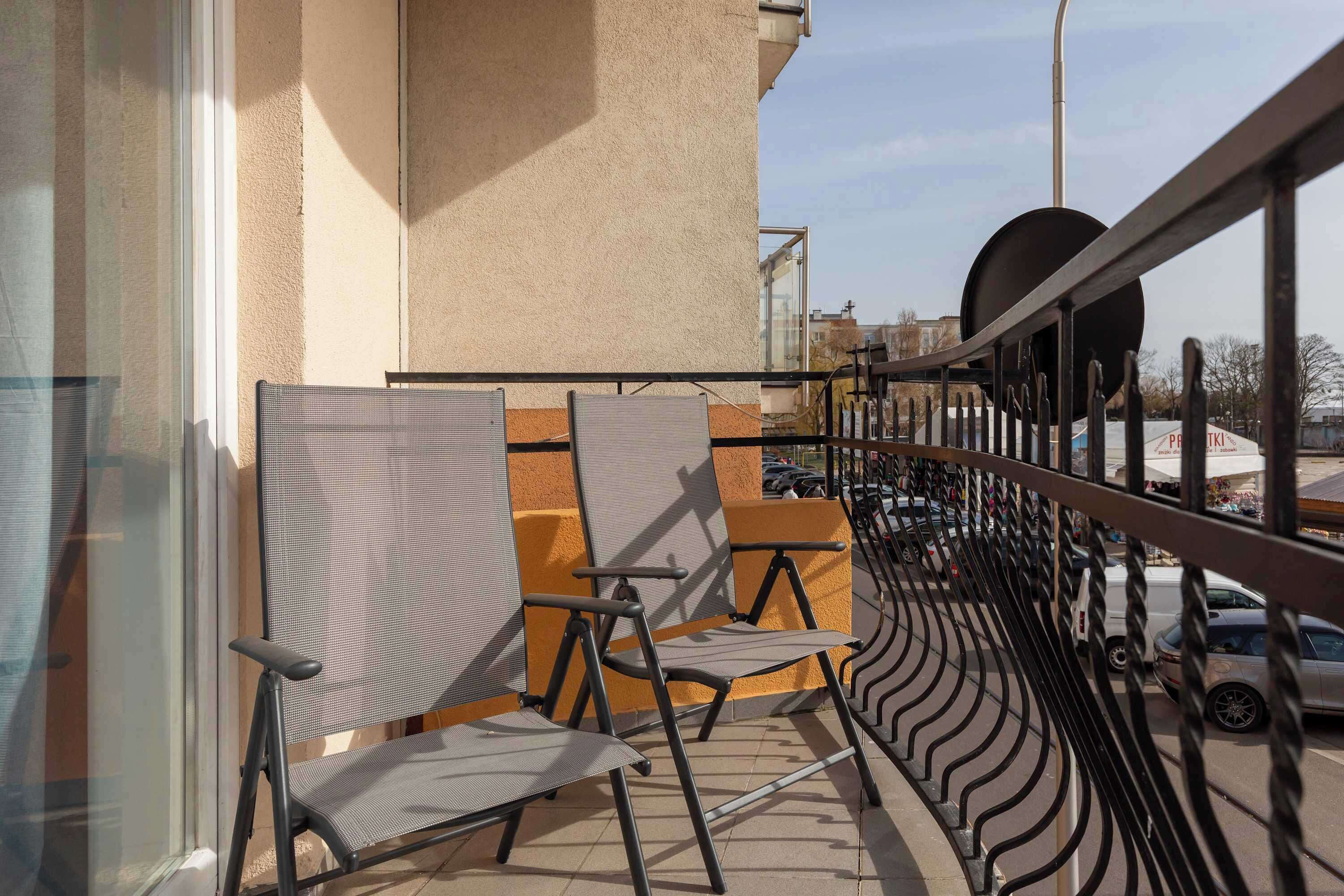 Ein Balkon mit zwei Stühlen, einer Brüstung und Blick auf geparkte Autos und Gebäude.