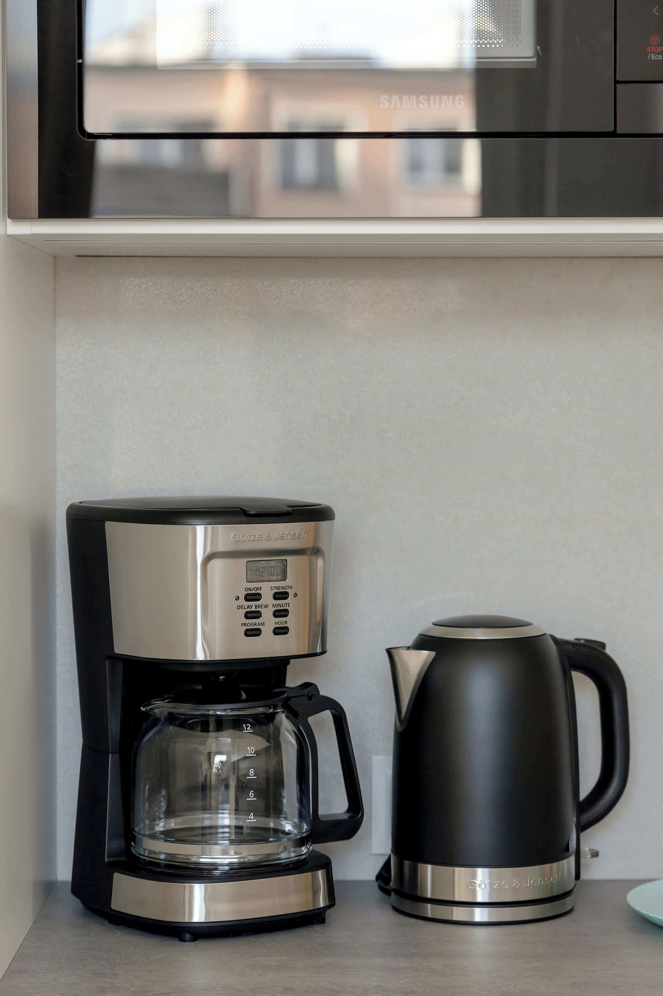 Eine kompakte Kaffeemaschine und ein Wasserkocher auf einer Arbeitsplatte, darüber eine Mikrowelle.