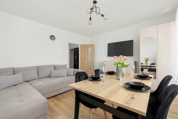 Ein modernes Wohnzimmer mit einem grauen Ecksofa, einem Holzesstisch mit schwarzen Tellern und einer Blumenvase, sowie einem wandmontierten Flachbildfernseher.