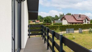 Ein Balkon mit Holzboden und Metallgeländer, der auf eine Rasenfläche und ein Haus mit rotem Dach blickt.
