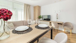 A modern dining area with a wooden table, two white plates, and wine glasses. There is a sofa and a TV in the background.