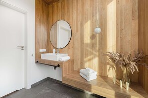 Ein modernes Badezimmer mit Holzwand, rundem Spiegel, weißem Waschbecken und Handtuchhalter.