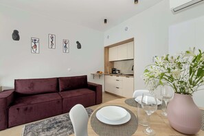 En moderne stue med en mørkerød sofa, et spisebord med hvide tallerkener og glas, samt en rosa vase med blomster.