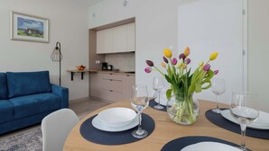 Un comedor con mesa de madera dispuesta para cuatro personas, un sofá azul, una cocina al fondo y un florero con flores sobre la mesa.