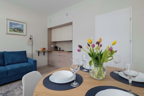 A dining area with a wooden table set for four, a blue sofa, a kitchen in the background, and a vase with flowers on the table.