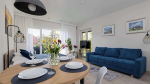 Una sala de estar moderna con una mesa de comedor redonda dispuesta para dos personas, un sofá azul y un televisor sobre un mueble bajo.