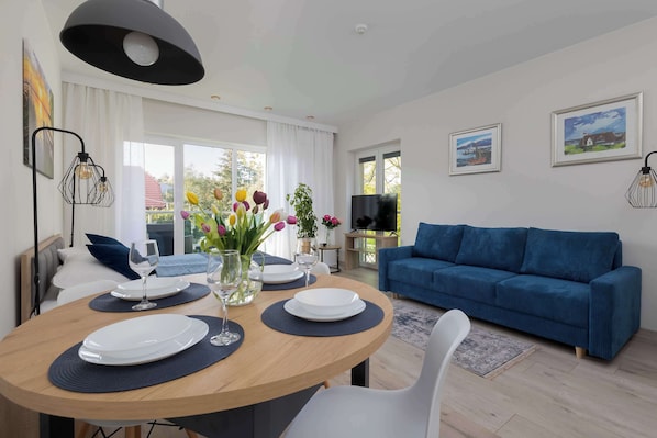 A modern living room with a round dining table set for two, a blue sofa, and a television on a stand.