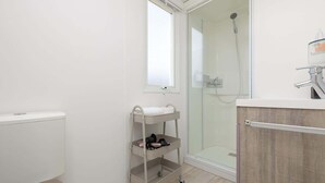 Un baño con inodoro, ducha, zona de ducha con mampara de vidrio, ventana, repisa con artículos de aseo y tocador de madera.