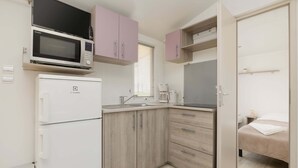 Una kitchenette compacta con microondas, refrigerador y lavabo. Hay una cama con una manta blanca y una mesita de noche pequeña.