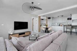 A modern living room with a large flat-screen TV, a grey sofa, and a dining area with a marble countertop and chairs.