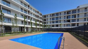 A swimming pool with a wooden deck, surrounded by a multi-story building with balconies and a fence.