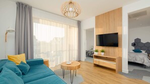 A modern living room with a sofa, a round wooden coffee table, a TV mounted on a wooden wall, and a bed with a geometric headboard.