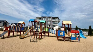 A beach playground with slides and climbing structures, set against a backdrop of modern buildings.