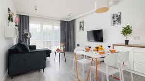 Una sala de estar moderna con un sofá negro, una mesa de comedor con sillas blancas, un televisor y una planta en maceta.
