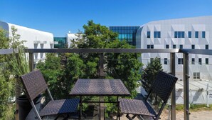 A balcony with two wicker chairs and a small table, overlooking a modern building and trees.