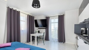 A modern hotel room with a large bed, a desk with a chair, a flat-screen TV, and a kitchenette.