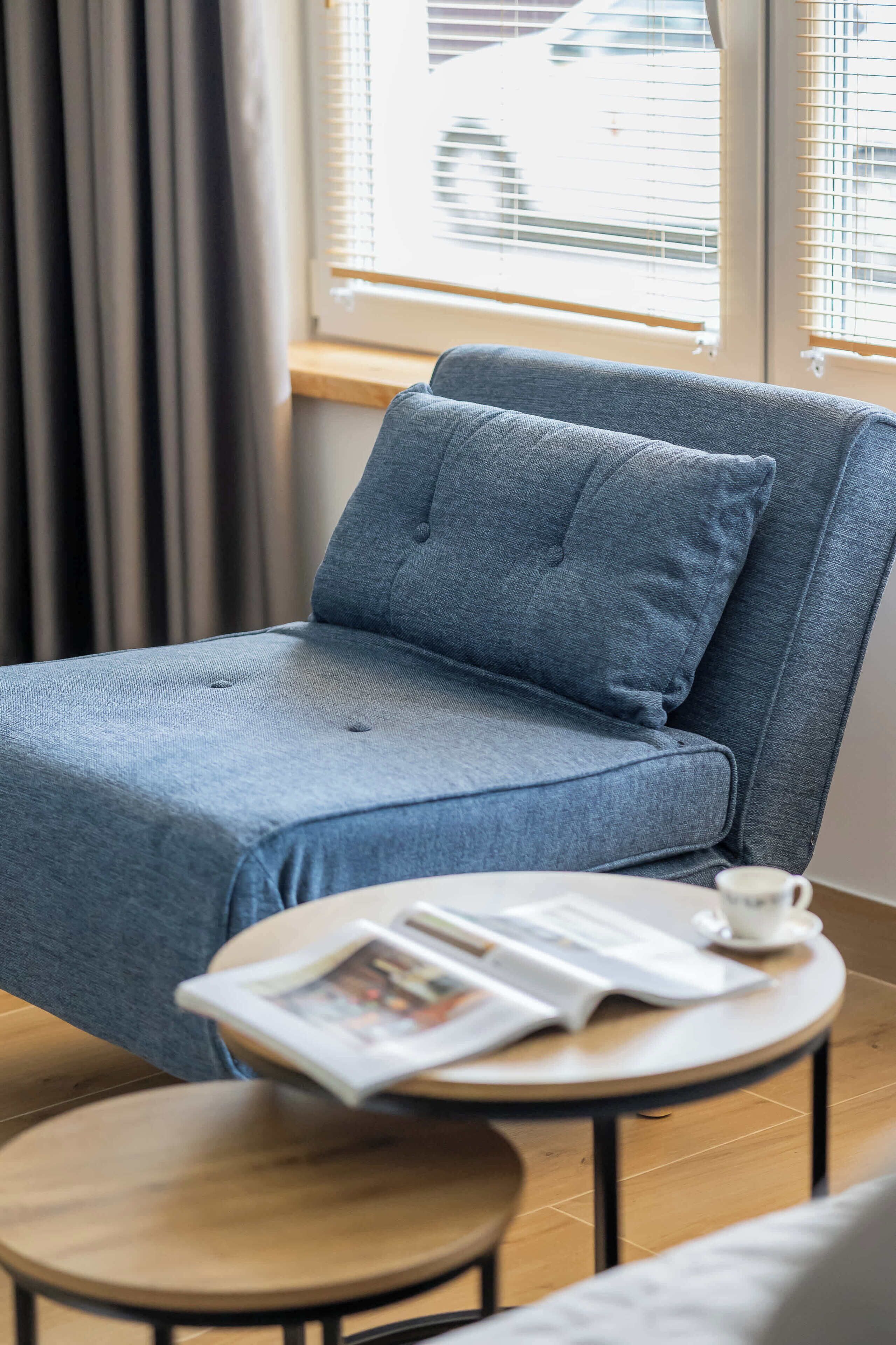 Une pièce avec un canapé gris, une petite table ronde sur laquelle se trouvent une tasse et un journal, et une fenêtre avec des stores.
