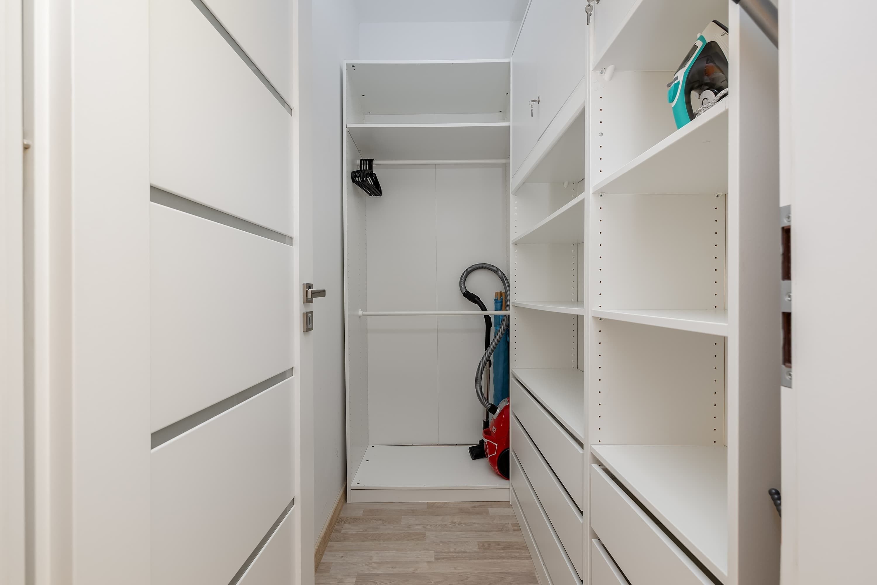 Un vestidor estrecho con estantes blancos, un aspirador y una puerta a la izquierda.
