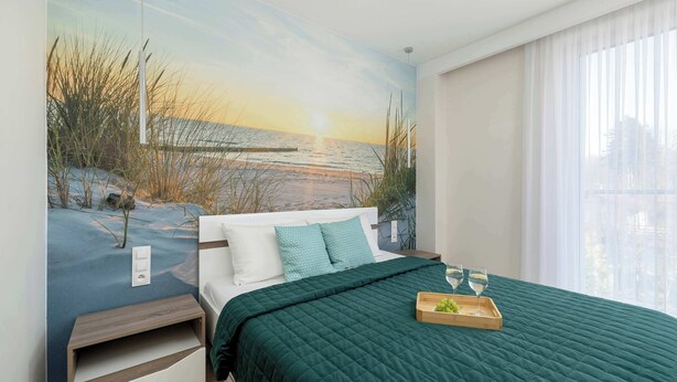 Une chambre avec une fresque murale représentant un coucher de soleil sur la plage, un lit recouvert d’une couverture verte, une table de chevet et un plateau avec des verres et des fruits.