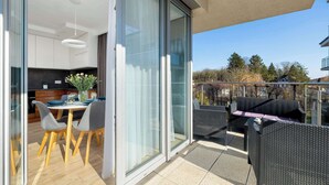 Un appartement moderne doté d’un balcon, d’un coin repas avec une table et des chaises, et d’une cuisine visible à travers des portes vitrées.