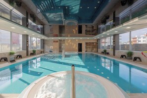 Une grande piscine intérieure avec un jacuzzi au centre, entourée de rampes de verre et de plantes en pot.