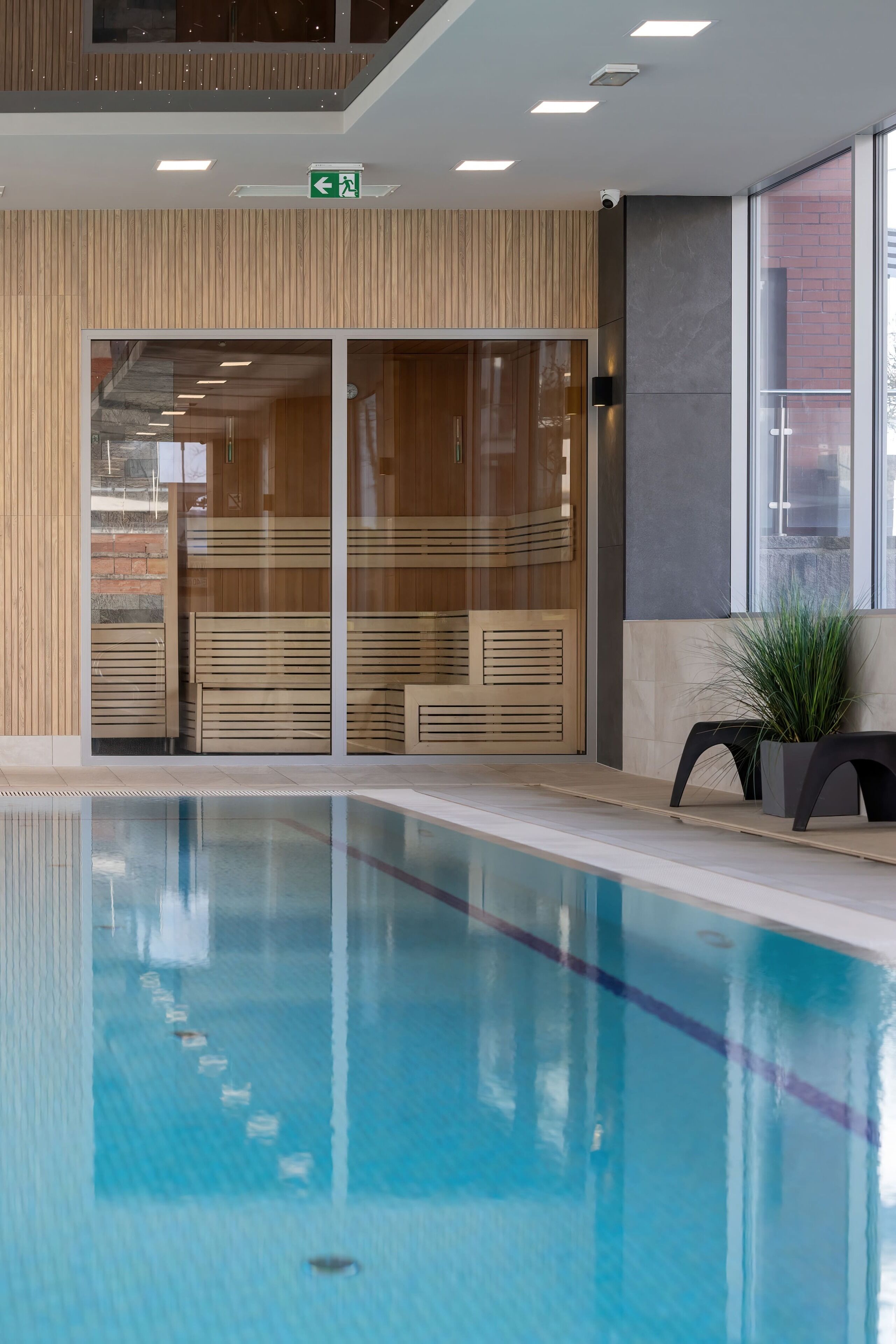 A modern indoor pool area with clear glass doors, wooden paneling, and a potted plant.
