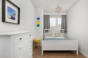Une chambre à coucher avec un lit blanc, un petit tabouret jaune, une commode et un tableau encadré accroché au mur.