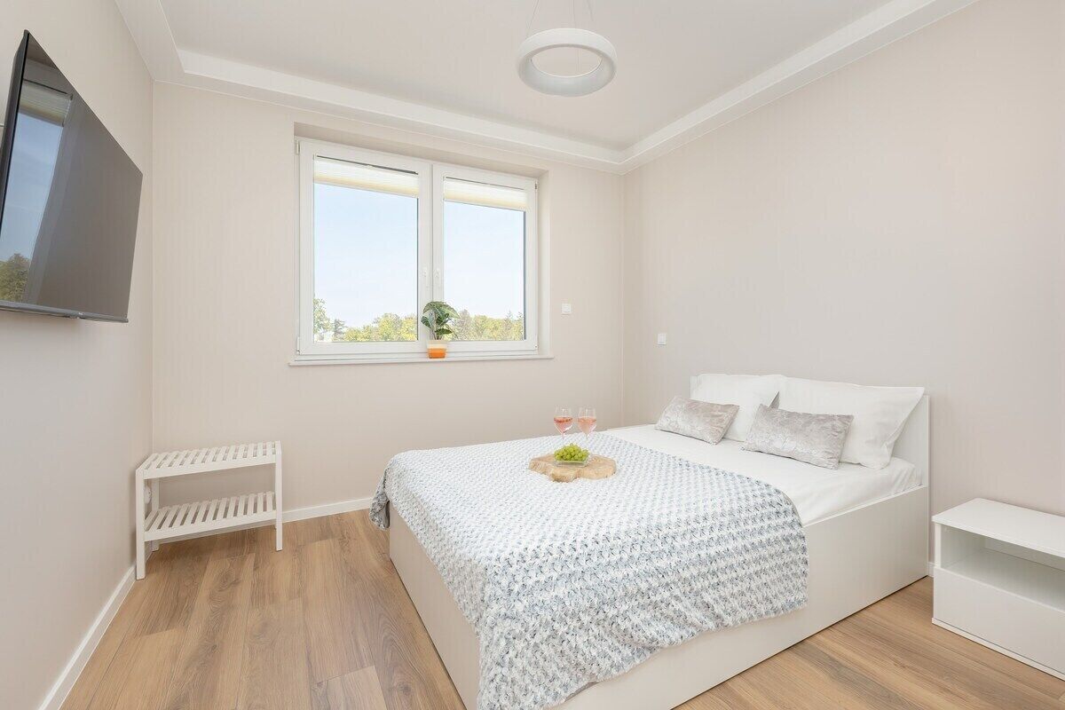 A modern bedroom with a bed, a TV, a window, and a small shelf.