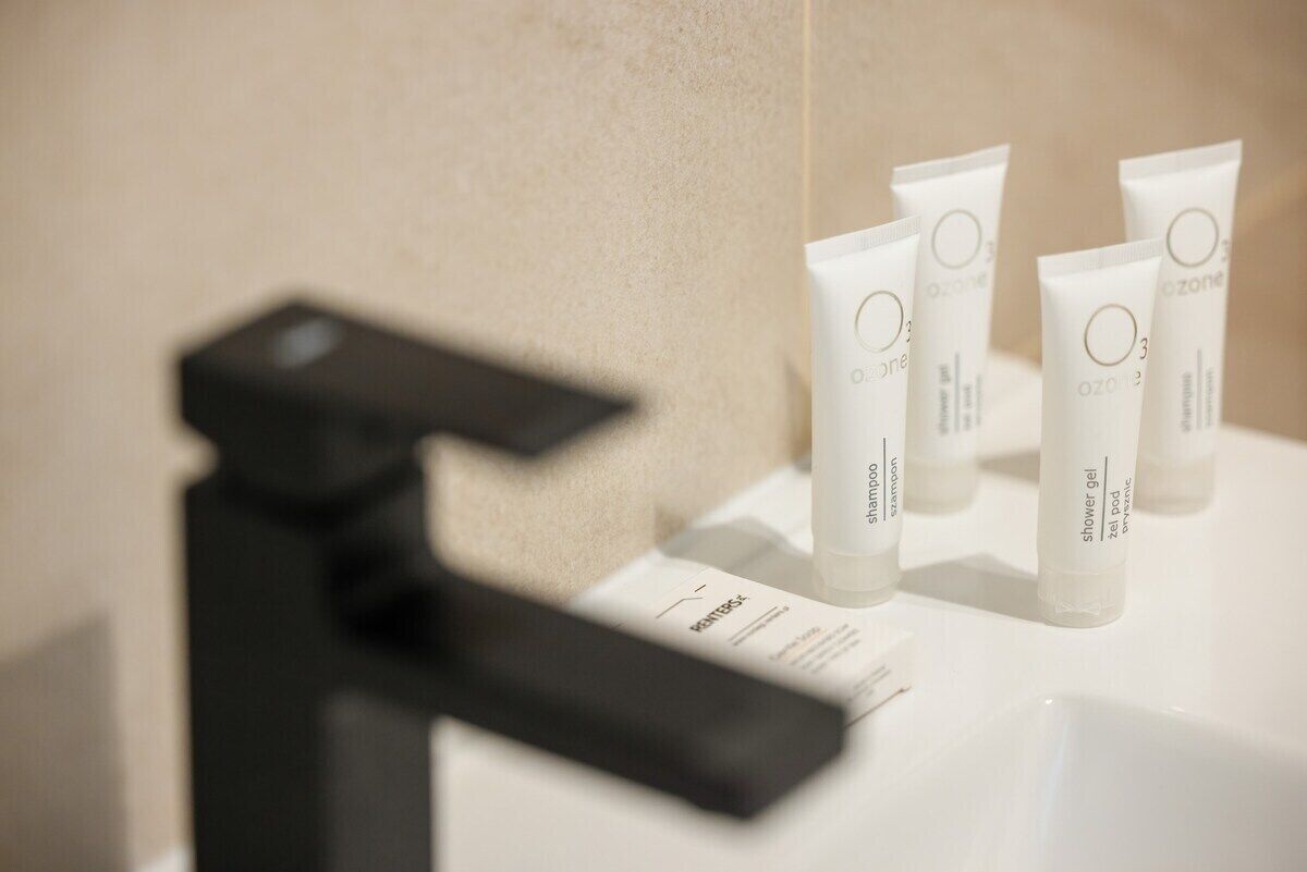 A bathroom with a black faucet, a white sink, and several tubes of Ozone shower gel and shampoo.