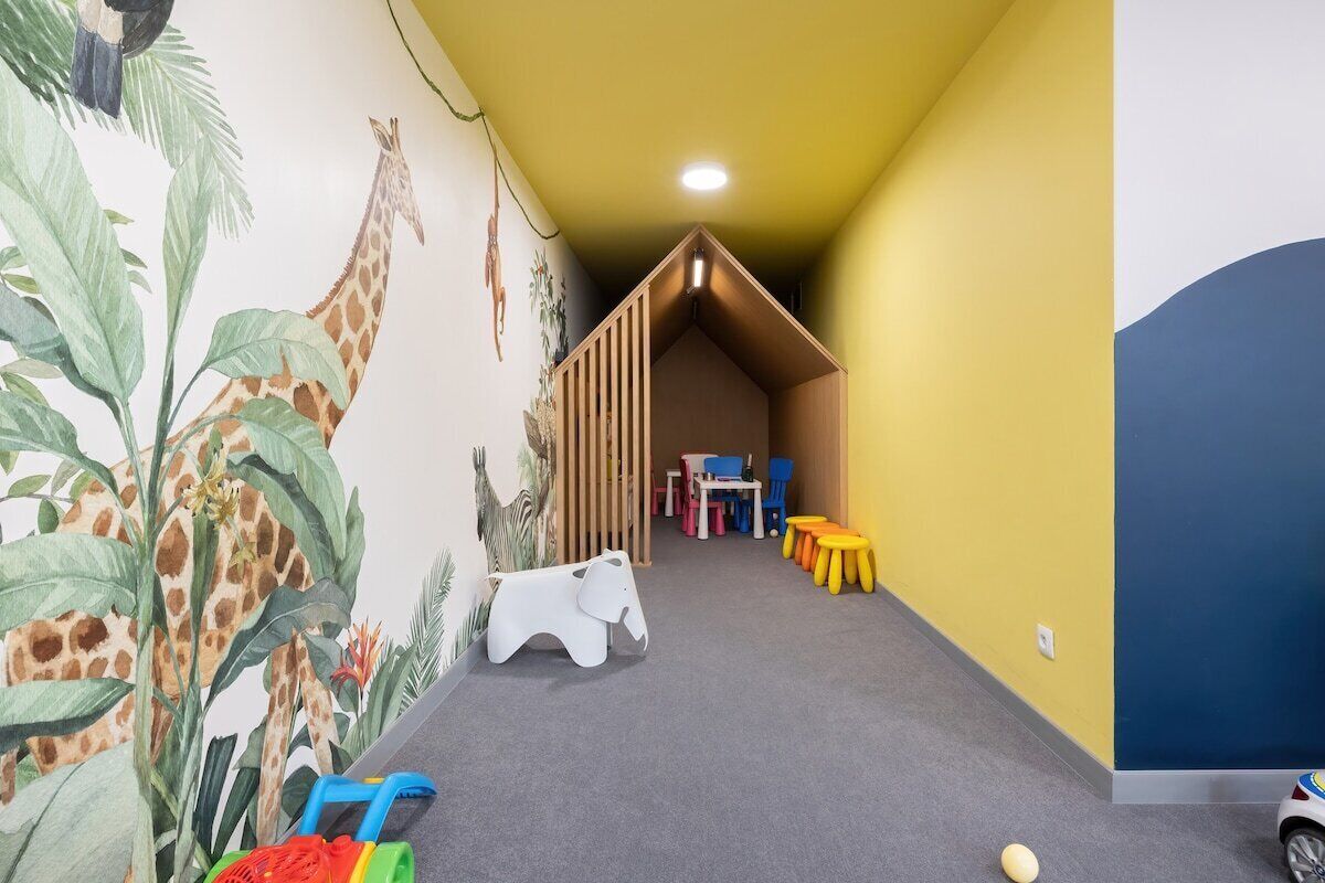 Un espace de jeux pour enfants agrémenté d’une fresque représentant des girafes et des éléphants, d’une maisonnette en bois et de tabourets colorés.
