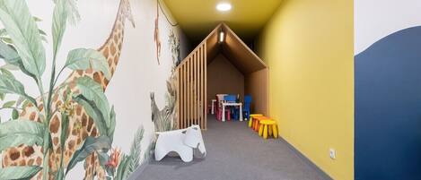 A children's play area with a mural of giraffes and elephants, a wooden playhouse, and colorful stools.