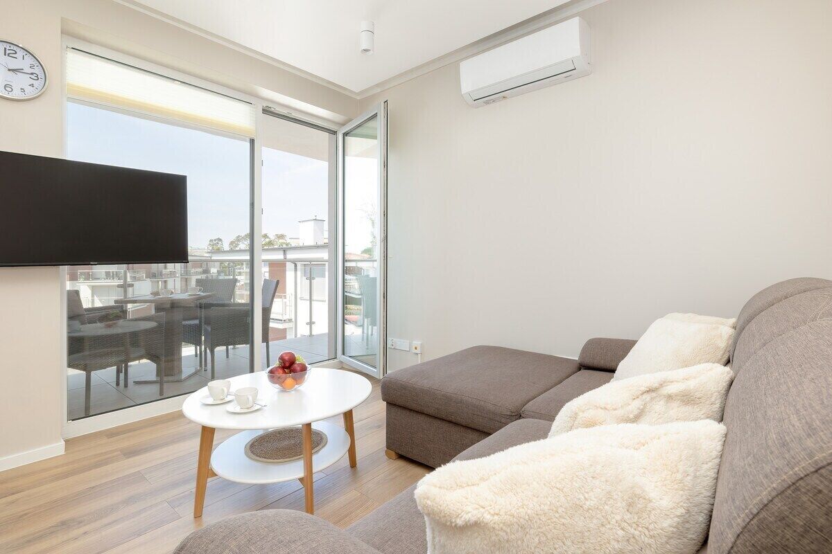 A modern living room with a sofa, a small round table with cups and fruit, a flat-screen TV, and a large window with a view of outdoor furniture.