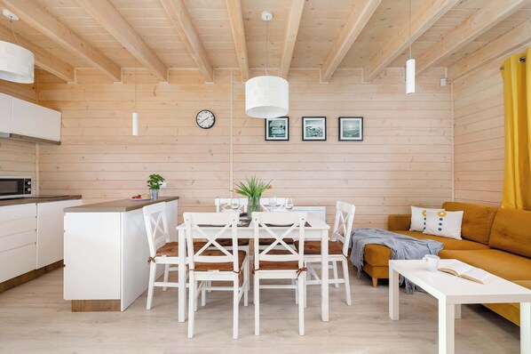 Een modern interieur met een houten plafond, een eethoek met witte stoelen en een tafel, een bank met gele kussens, en een keuken met witte kastjes.
