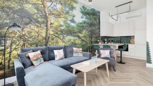 A modern living room with a large sofa, a coffee table, and a view of a forest through a mural. - Spacious Apartment | Close to the Beach, Parking. (Mrzeżyno)