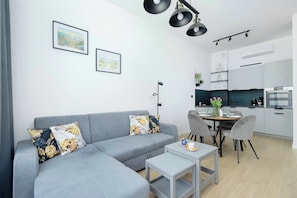 En moderne stue med en grå sofa, et lille bord og et spiseområde med stole. Væggen er pyntet med billeder, der er en loftlampe, og et køkken ses i baggrunden.