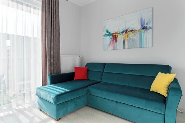 A teal sectional sofa with red and yellow cushions, a large abstract painting on the wall, and a view through sheer curtains.
