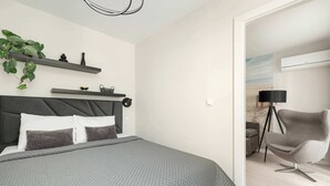 A modern bedroom with a bed, grey bedding, a headboard, a bedside table with a lamp, and a potted plant.