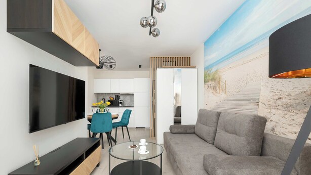 A modern living room with a flat-screen TV, a glass coffee table, a grey sofa, and a kitchen in the background.