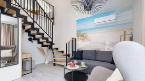 Una sala moderna con un sofá gris, una mesa de centro redonda con frutas, una escalera con barandillas negras y un mural de playa en la pared.