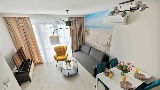 Un salon moderne avec un canapé, une table à manger dressée pour quatre personnes et une vue sur la plage, visible à travers une grande fenêtre.