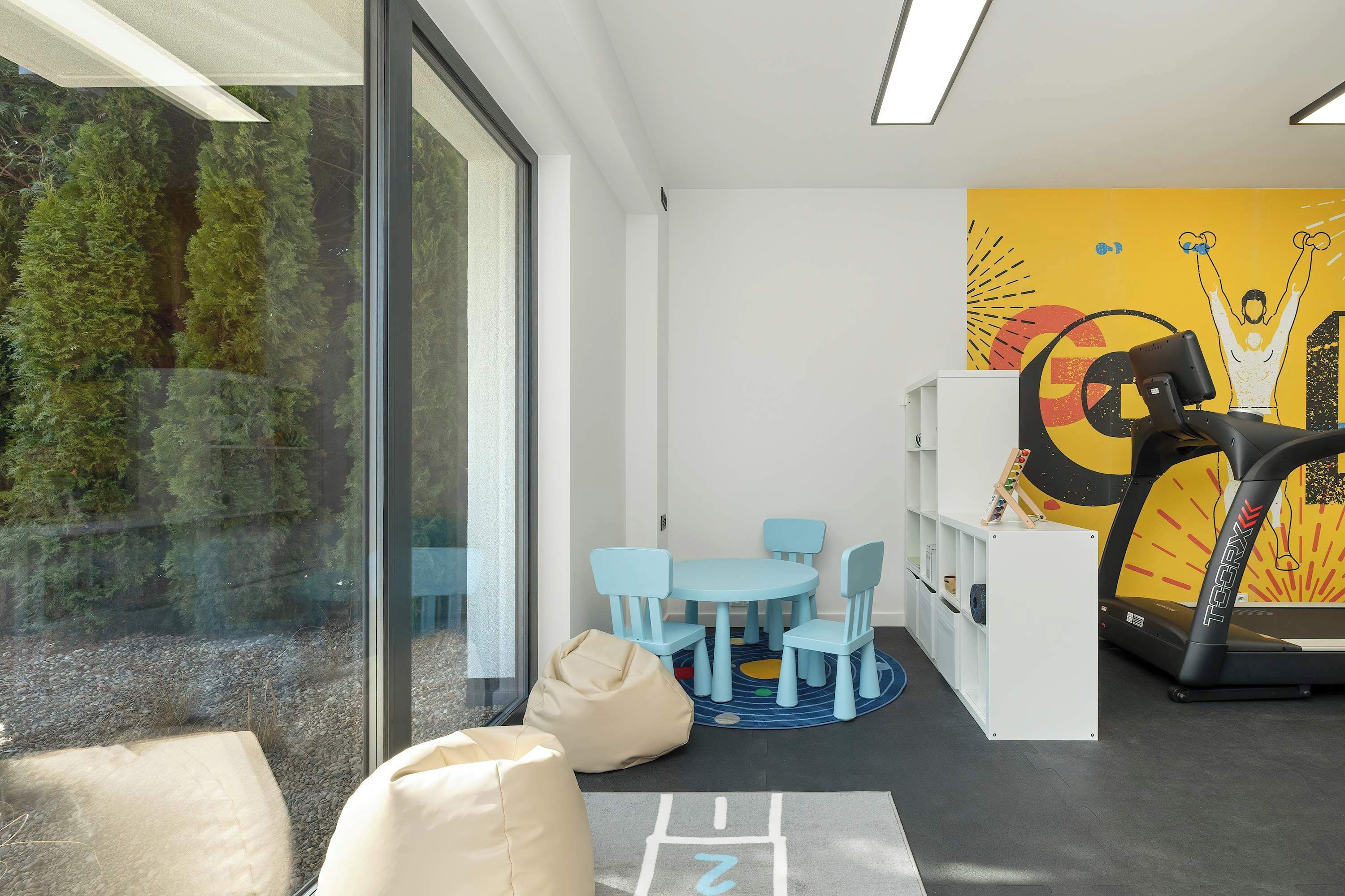 A modern room with a large window, a treadmill, a blue table with chairs, and a bookshelf.