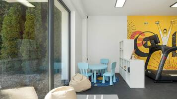 A modern room with a large window, a treadmill, a blue table with chairs, and a bookshelf.