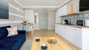 Una sala de estar moderna con un sofá azul, una mesa de centro de madera con un frutero y una cocina con armarios blancos y un microondas.