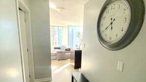 En hall med en klocka på väggen, en bänk och utsikt över ett vardagsrum med en soffa och ett matbord.