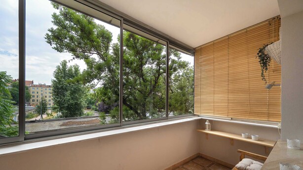 Ein Balkon mit großem Fenster, das auf Bäume und Gebäude blickt, einer Holzbank und einem kleinen Regal mit Gegenständen.