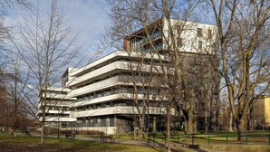 A multi-story building with a modern architectural design, surrounded by a park with benches and trees.