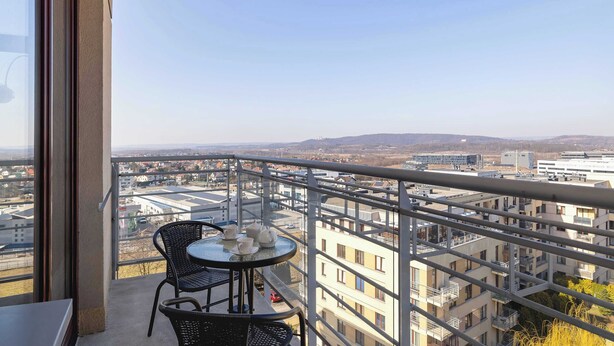 A balcony with a table and chairs, overlooking a cityscape with buildings and distant hills.