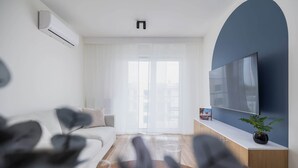 A modern living room with a white sofa, a flat-screen TV, and a large window with sheer curtains.